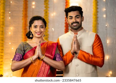 Happy young indian couple in traditional ethnic clothing saying namaste or welcome gesture by looking camera during festival celebration at home - concept of greeting, wishes and togetherness.