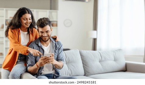 Happy young indian couple using smartphone, wife pointing at cellphone screen, sitting on sofa in living room interior at home. New app ad, banner - Powered by Shutterstock