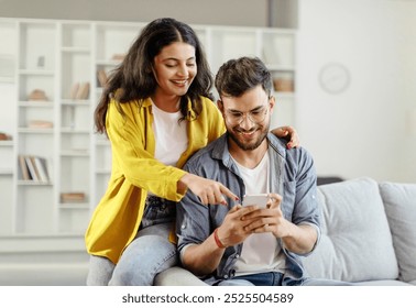 Happy young indian couple using smartphone, wife pointing at cellphone screen, sitting on sofa in living room interior at home. New app ad, banner - Powered by Shutterstock