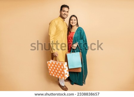 Happy young indian couple wearing traditional cloths holding shopping bags standing isolated on beige studio background. Diwali celebration and festive sale, copy space.