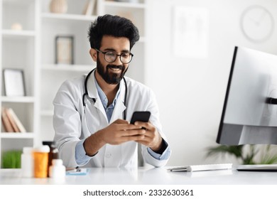 Happy young indian doctor in glasses and white coat sitting at table, using cellphone applications in clinic office. Smiling handsome general practitioner giving online mobile consultation, copy space - Powered by Shutterstock