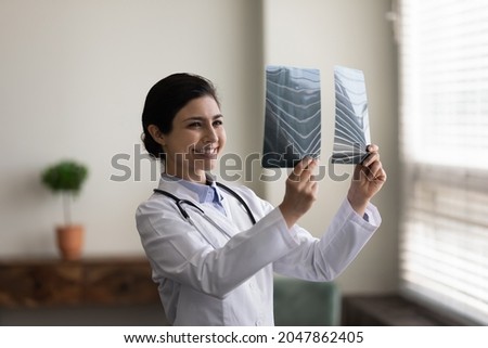 Happy young Indian doctor looking at xray radiography images. Physician, surgeon reviewing scan of patient bones, smiling at good screening test result. Medical checkup, healthcare, radiology concept