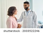 Happy young Indian doctor therapist in white coat has appointment consulting supporting putting hand on shoulder of older senior female patient in modern clinic hospital. Medical healthcare concept.