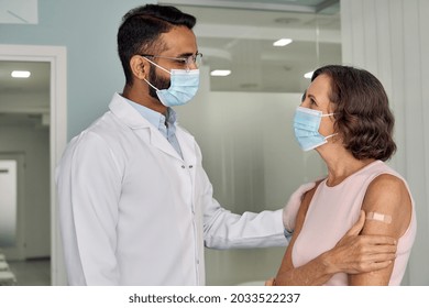 Happy Young Indian Doctor Therapist In White Coat Wearing Face Mask Supporting Older Senior Female Patient In Modern Clinic Hospital After Injection. Vaccination Coronavirus Safety Concept.