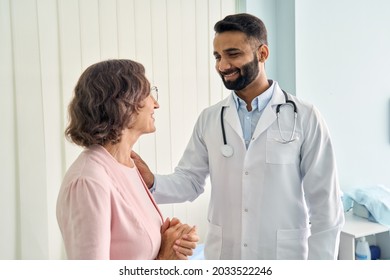 Happy young Indian doctor therapist in white coat has appointment consulting supporting putting hand on shoulder of older senior female patient in modern clinic hospital. Medical healthcare concept.