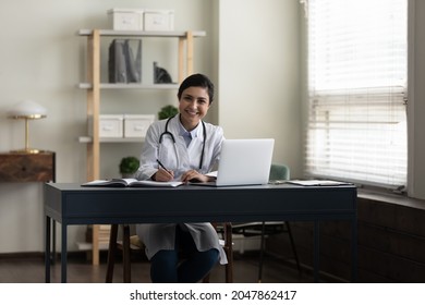 Happy young Indian doctor, therapist, general practitioner at work table with laptop in office, looking at camera, smiling, writing notes, medical records. Medic care concept. Professional portrait - Powered by Shutterstock