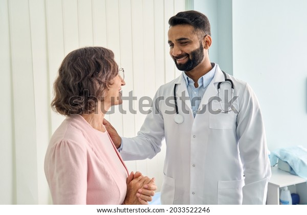 Happy young Indian doctor therapist in white\
coat has appointment consulting supporting putting hand on shoulder\
of older senior female patient in modern clinic hospital. Medical\
healthcare concept.