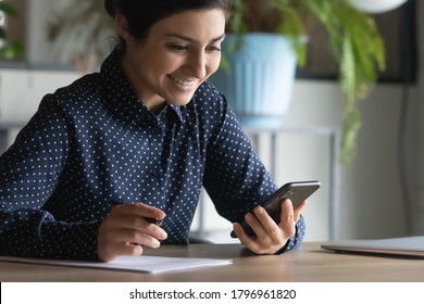 Happy young indian ethnicity female manager looking at smartphone screen, web surfing important information while preparing paper report, smiling mixed race student writing essay, using internet.