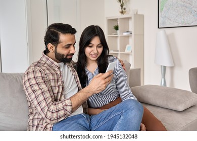 Happy Young Indian Family Couple Using Smart Phone Looking At Cellphone Sit On Sofa Together Relaxing At Home. Smiling Husband And Wife Checking Social Media Apps Or Shopping In Mobile Application.