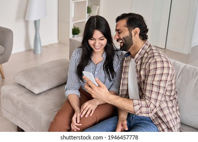 Happy Young Indian Family Couple Laughing Using Smart Phone Looking At Cellphone Sit On Sofa Together At Home. Smiling Husband Showing Wife Funny Video In Application Having Fun With Mobile Device.