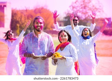 Happy young indian family wearing white kurta and holding plate full of laddu sweet and gula celebrating holi festival wtih colorful people at park outdoor.