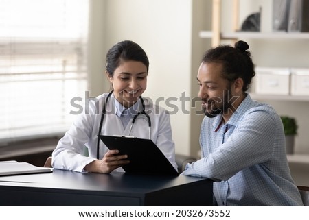 Happy young indian female gp doctor showing test result on clipboard to interested millennial african ethnicity male patient, discussing healthcare medical insurance or illness treatment at clinic.