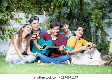 Happy Young Indian Friends Sitting At Park Or Garden Playing Guitar And Music Having Fun Together And Enjoy Outdoor Picnic.