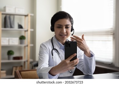 Happy young indian general practitioner therapist in headset holding video call web camera conversation on cellphone, communicating distantly with patient or e-learning on virtual conference event. - Shutterstock ID 2032673723