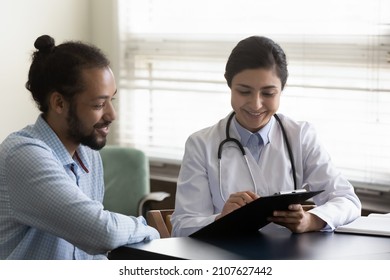 Happy young Indian general practitioner doctor showing health test result on clipboard to African American male patient, discussing medical insurance agreement or analyzing disease history at clinic.