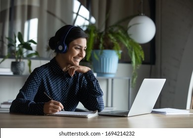 Happy young indian girl with wireless headphones looking at laptop screen, reading listening online courses, studying remotely from home due to pandemic corona virus world outbreak, quarantine time.