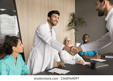 Happy young Indian male doctor giving handshake to cooperation partner, grateful patient on medical business meeting, smiling, laughing. Positive clinic staff discussing healthcare service - Powered by Shutterstock