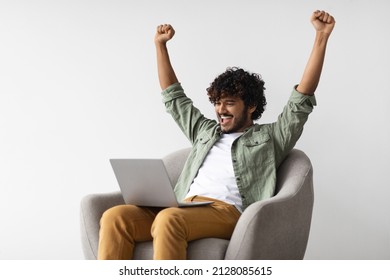 Happy Young Indian Man In Casual Sitting In Armchair With Computer On His Lap, Looking At Laptop Screen And Raising Hands Up, Unemployed Guy Got New Job, Copy Space. Job Search Websites Concept