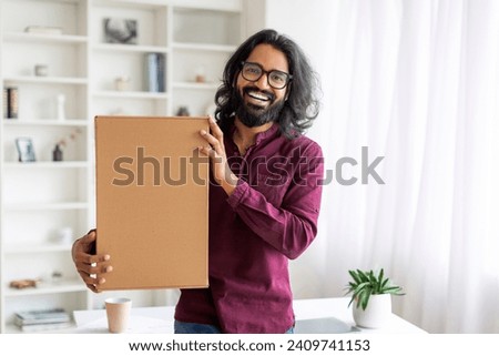 Happy Young Indian Man Embracing Cardboard Box With Delivery At Home, Handsome Smiling Eastern Male Holding Delivered Parcel In Hands While Standing Near Desk In Living Room, Copy Space