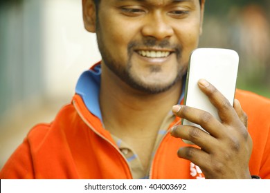 Happy Young Indian Man Smiling As He Looks On At His Smartphone Using Internet With Broadband Wifi And 4g Connected - Concept Of Asian People Making Use Of Technology To Communicate With Family