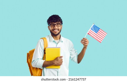 Happy Young Indian Man Studying At American University. Cheerful South Asian Exchange Program Student In Glasses With Books And Bag Standing On Blue Studio Background, Holding Flag Of USA And Smiling