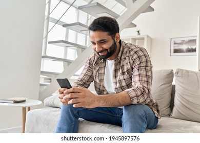 Happy Young Indian Man Using Smart Phone Sitting On Couch At Home. Smiling Bearded Ethnic Guy Holding Mobile Phone Watching Social Media Video On Smartphone, Ordering Food Delivery Online Or Messaging