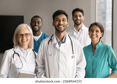 Happy young Indian medical boss and diverse team of doctors, practitioners, surgeons in uniforms looking at camera with toothy smiles, posing for portrait. Multiethnic small clinic staff shot - Powered by Shutterstock