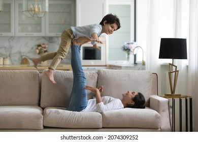 Happy young Indian mom having fun with little son at home, playing active games in couch, lying on sofa, holding boy, supporting kid on legs and feet for kid making airplane with open flying hands