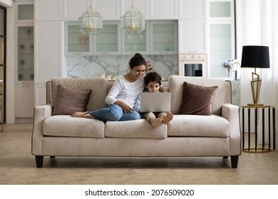 Happy Young Indian Mother And Excited Little Son Playing Virtual Online Videogame On Laptop In Living Room. Mom And Kid Making Video Call On Computer, Resting On Couch In Pale Home Interior. Wide Shot