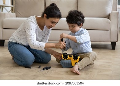 Happy young Indian mother helping adorable small kid son fixing toy car with screwdriver, playing together at home. Smiling mum teaching little child repairing, developing motility skills