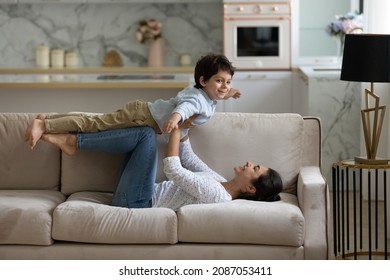 Happy young Indian mother lifting in air on straight arms joyful cute small child son, lying on cozy couch. Joyful family practicing balance doing yoga exercises, having fun playing at home.