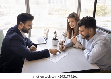 Happy young Indian realtor man pointing finger at document for cheerful husband and wife signing, giving legal consultation, support. Positive couple setting signature in loan agreement - Powered by Shutterstock