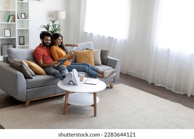 Happy Young Indian Spouses Relaxing With Modern Gadgets On Couch At Home, Married Eastern Couple Resting In Living Room Interior, Man Using Digital Tablet While Woman Browsing App On Smartphone - Powered by Shutterstock