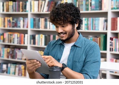 Happy young indian student in earphones using mobile tablet sitting in library over bookshelves. Teenage guy enjoy reading ebook on digital device watching education webinar, training video tutorial - Shutterstock ID 2071252031