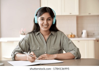 Happy Young Indian Teen Girl Woman Wearing Headphones Looking At Camera Studying Listening Audio Course Remote Learning English Language Writing Notes Working Sitting At Table At Home Office. Portrait