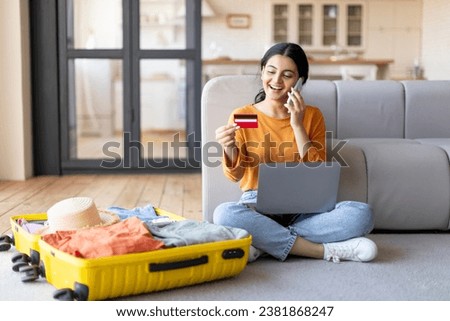 Happy young indian woman with credit card and cellphone booking vacation at travel agency or making hotel reservation online, using laptop computer, planning trip, sitting next to suitcase at home