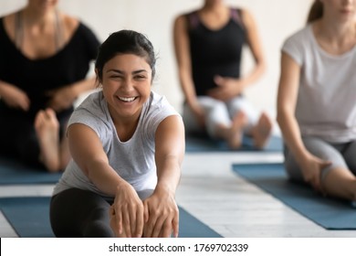 Happy young indian woman excited by flexibility progress in seated forward bend position, enjoying group yoga class indoors. Smiling beautiful fit biracial lady practicing Paschimottanasana in studio.