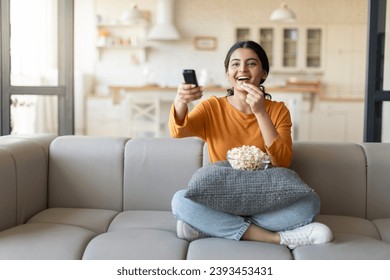 Happy Young Indian Woman Holding Remote Controller And Eating Popcorn At Home, Cheerful Millennial Eastern Lady Watching TV Show While Relaxing On Couch In Cozy Living Room, Copy Space - Powered by Shutterstock