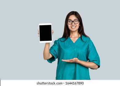 Happy Young Indian Woman Professional Nurse Female Doctor Wear Green Uniform Show Digital Tablet Mock Up Blank Empty Screen Advertise Medical Care App Tech Concept Isolated On Gray Studio Background.