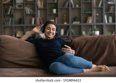 Happy young Indian woman relax on sofa in living room wear headphones listen to music on smartphone. Smiling millennial ethnic female rest at home enjoy good quality sound in earphones use cellphone.