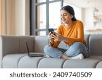 Happy young Indian woman using smartphone, sitting on sofa with laptop beside her, smiling eastern female enjoying a relaxed day at home, messaging on mobile phone with friends, copy space