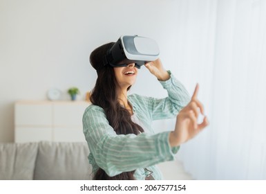 Happy Young Indian Woman In Virtual Reality Glasses Exploring Cyberspace, Touching Imaginary Screen, Playing Interactive Video Game At Home. Futuristic Technologies And Entertainments Concept