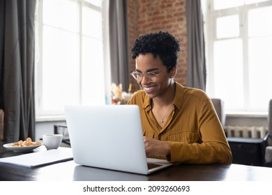Happy young inspired African American woman in eyeglasses looking at computer screen, reading email with good news, enjoying studying or working distantly on online project at modern home office.