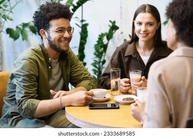Happy young intercultural couple having coffee and listening to friend sitting in front of them during discussion of funny stuff in cafe