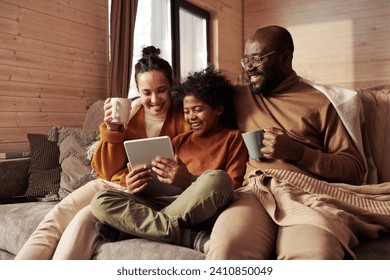 Happy young intercultural family of three looking at screen of tablet held by cute boy while sitting on couch and watching sitcom at leisure - Powered by Shutterstock