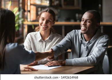 Happy young interracial couple shake hand of bank manager broker buy insurance services take mortgage loan, mixed ethnicity customers handshake agent make agreement financial business deal at meeting