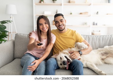 Happy Young Interracial Couple Sitting On Couch With Their Funny Dog And Watching TV At Home. Arab Man And His Caucasian Girlfriend With Pet Enjoying Television Program Indoors