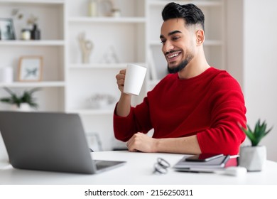 Happy Young Islamic Man With Beard Drinks Cup Of Tea At Workplace With Computer In Home Office Interior. Business, Coffee Break From Work, Pause And Vitality For Freelance During Covid-19 Outbreak