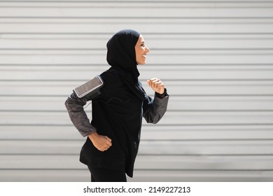 Happy young islamic woman athlete in hijab, sportswear with smartphone runs on wall background, profile. Jogging in morning, technology for training, sports in city, health care due covid-19 pandemic - Powered by Shutterstock