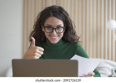 Happy young Italian female employee in glasses talks online on laptop with colleagues or business partner, showing thumb up, cheerful Spanish woman freelancer making video call while working remotely